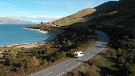 Slowmo-Aerial-Drone-following-a-campervan-with-a-view-of-beautiful-blue-Lake-Hawea,-field-of-sheep-and-mountains-in-late-afternoon-in-New-Zealand