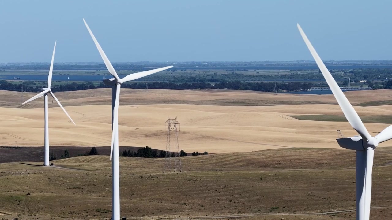 Aerial view of a wind farm with massive turbines spinning and generating clean, renewable energy against a backdrop of golden rolling hills and a clear blue sky in Rio Vista, California