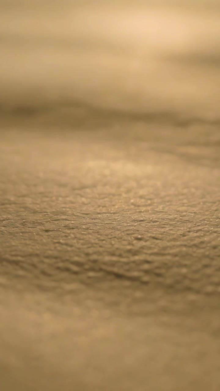 Vertical video: Moving camera shifting focus showing tan surface texture under warm low-angle light