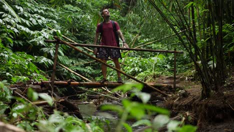 un joven con camiseta y pantalones cortos camina sobre un pequeño puente de bambú sobre un pequeño arroyo de agua, mira los hermosos alrededores y continúa cruzando