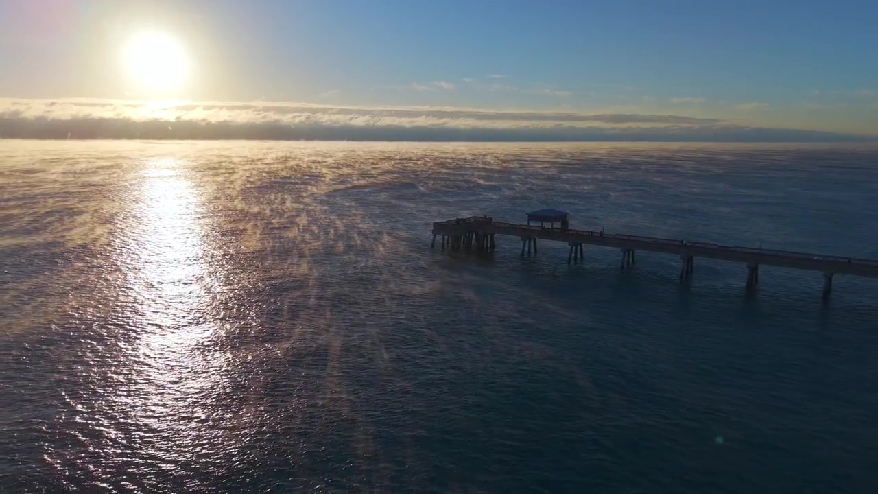 Imagem de drone em recuo revelando o píer de Deerfield Beach numa manhã fria ao nascer do sol com vapor subindo da superfície do oceano