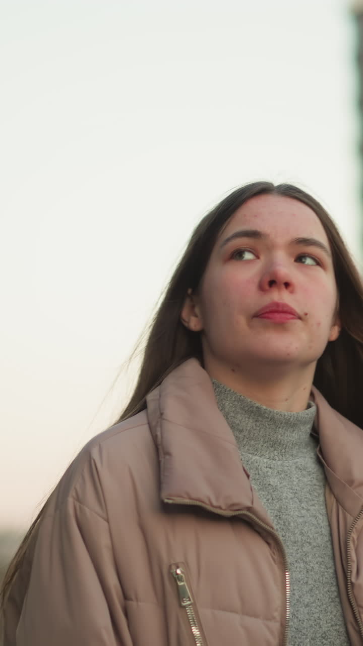una toma frontal de cerca de una joven con una chaqueta de melocotón y un suéter interior gris, capturada mientras camina por un parque urbano. la toma se centra en su parte superior del cuerpo y su expresión contemplativa