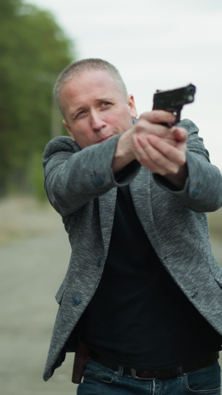 una vista cercana de un hombre con una chaqueta gris y pantalones vaqueros caminando por un camino de tierra al lado de una vía ferroviaria, sosteniendo una pistola apuntando con ella, con una vista borrosa de un bosque y líneas eléctricas lejanas