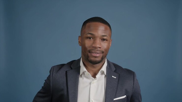 Telecamera che si abbassa su un uomo seduto che sorride in studio, mentre si prepara per un'intervista, indossando una spilla quadrata sull'abito.