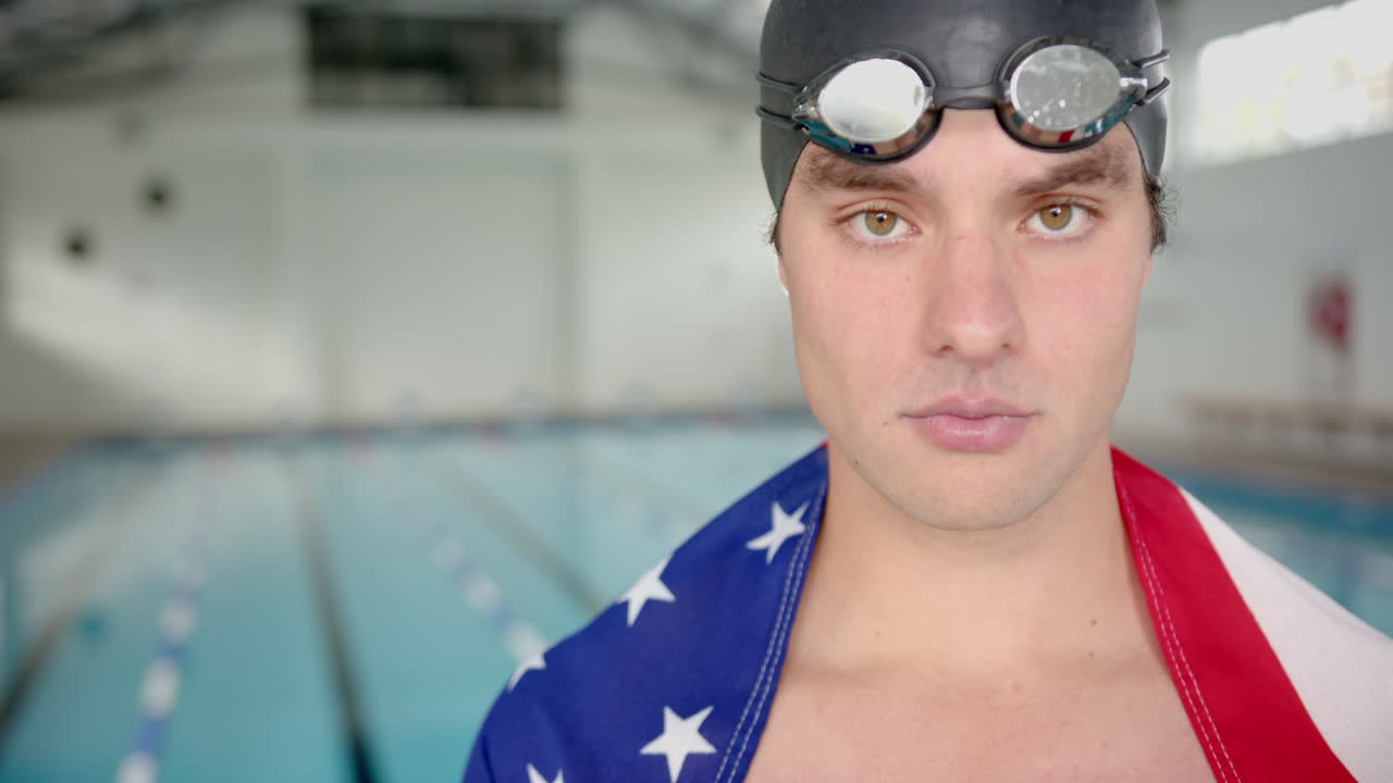 Male Swimmer wearing goggles and flag towel standing by indoor pool, looking determined, copy space