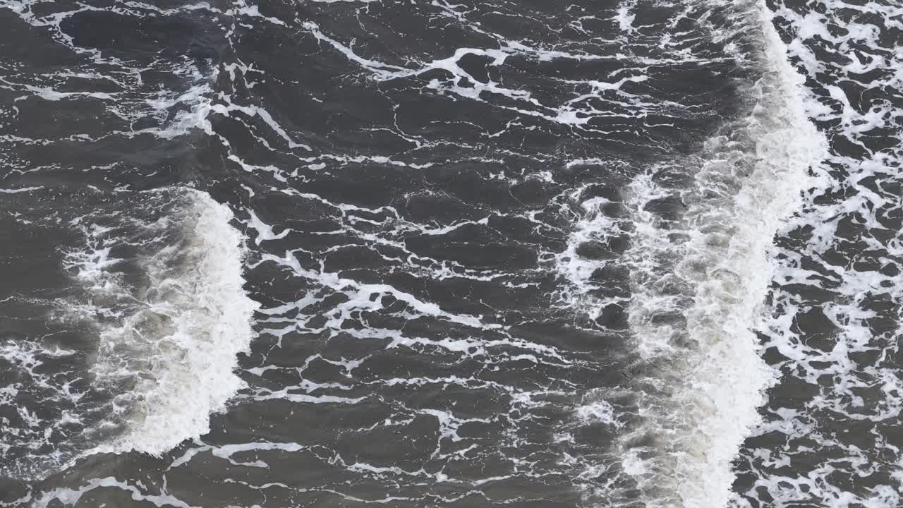 Overhead footage captures brown, sediment-laden ocean waves rolling and breaking along the shoreline in natural daylight, with steady camera movement and textured water patterns