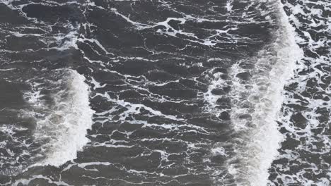 overhead footage captures brown, sediment-laden ocean waves rolling and breaking along the shoreline in natural daylight, with steady camera movement and textured water patterns