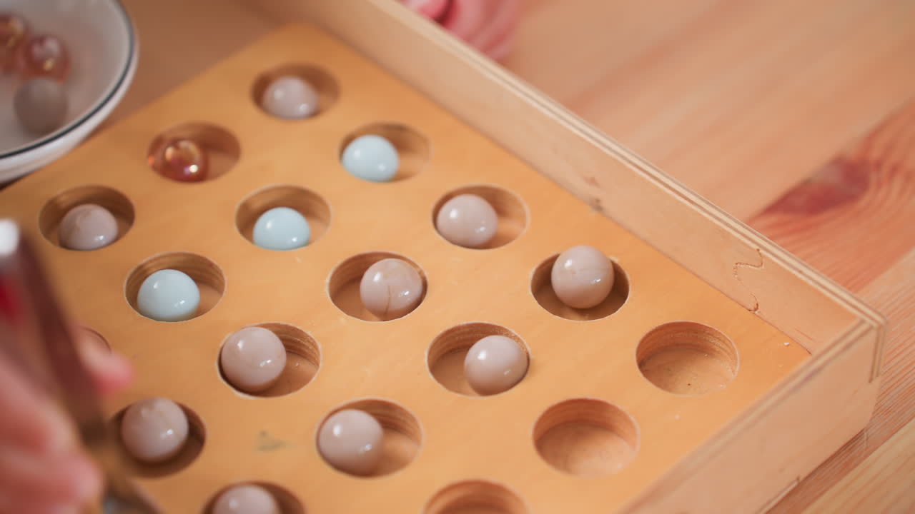 Close up hand view of toddler in checkered shirt carefully placing small balls into wooden puzzle box using tool, focusing on coordination and learning activity on smooth wooden table indoors
