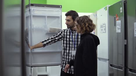 Young-woman-standing-in-front-the-open-door-of-refrigerator-with-male-consultant-discussing-design-and-quality-before-buying-in-a-consumer-electronics-store.-Discussing-characteristics-with-store-advisor