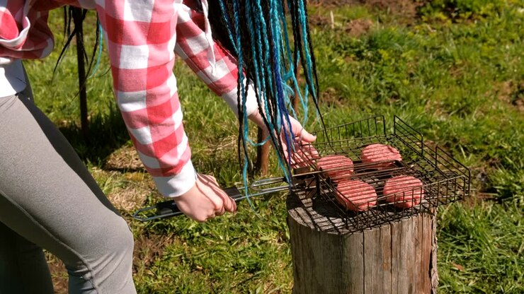Unrecognizable young woman closes the grill with the chops. Close-up hands. She is in plaid pink shirt and with blue braid hairs.