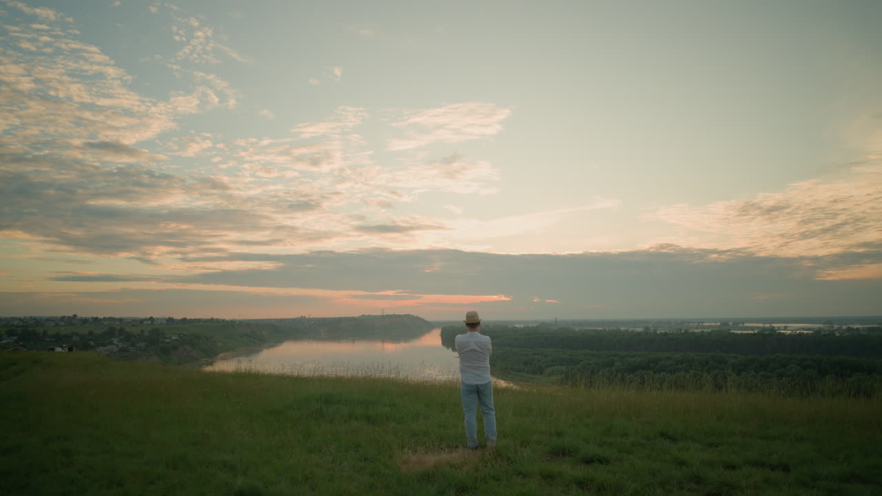A man stands contemplatively in a grassy field by a tranquil lake at sunset. He is dressed in a white shirt, hat, and jeans, capturing a peaceful, reflective moment as the sun sets on the horizon