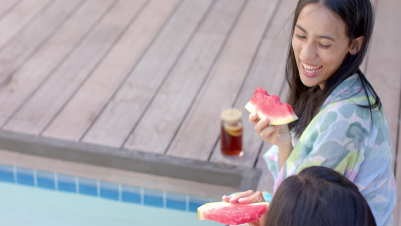Two biracial teenage girls enjoy watermelon by pool, copy space