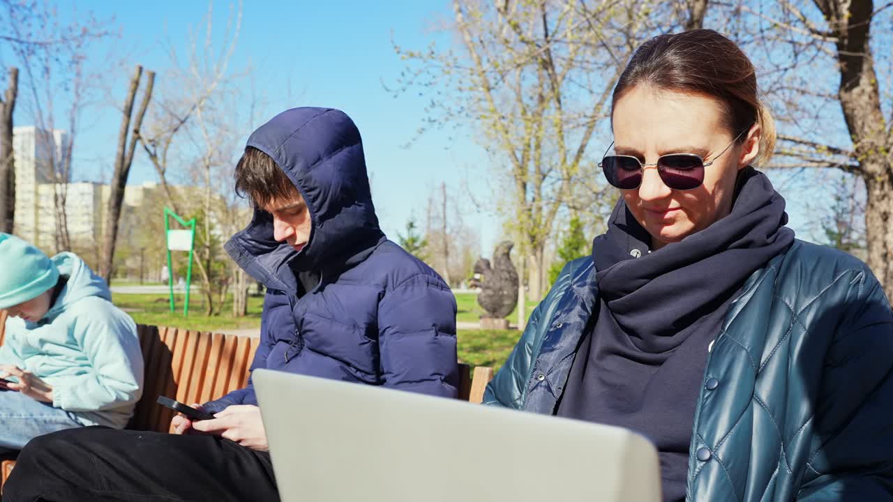 Kvinde og teenagere bruger digitale enheder på en parkbænk