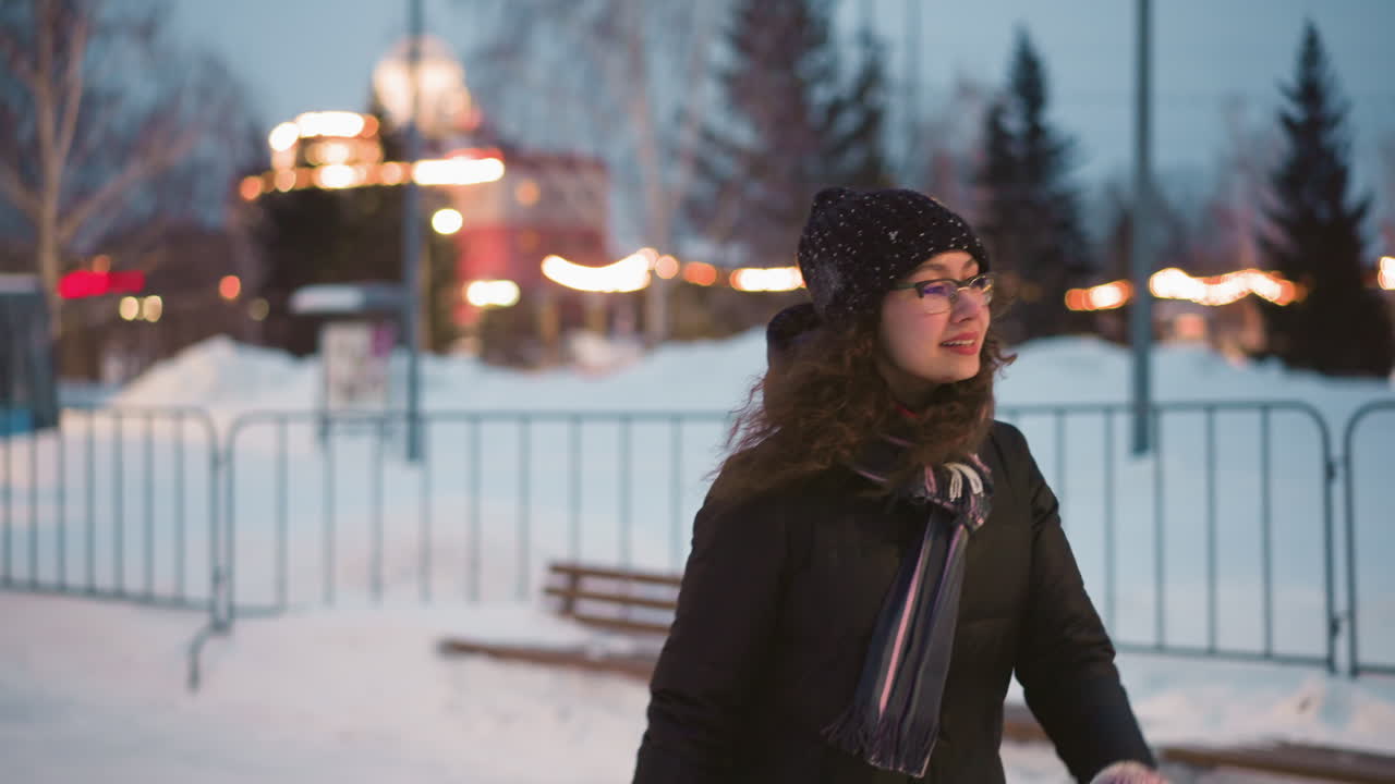 Skater with curly hair wearing glasses, black hat, and scarf walks through snowy outdoor area at dusk, enjoying cold winter , warm dressed in jacket, smiling while surrounded by festive blurred city lights
