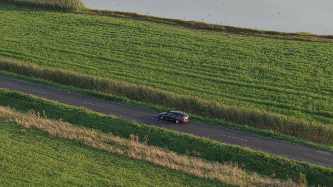 A dark car travels along a quiet asphalt road surrounded by lush green fields. The aerial view captures the peaceful countryside atmosphere in warm evening light