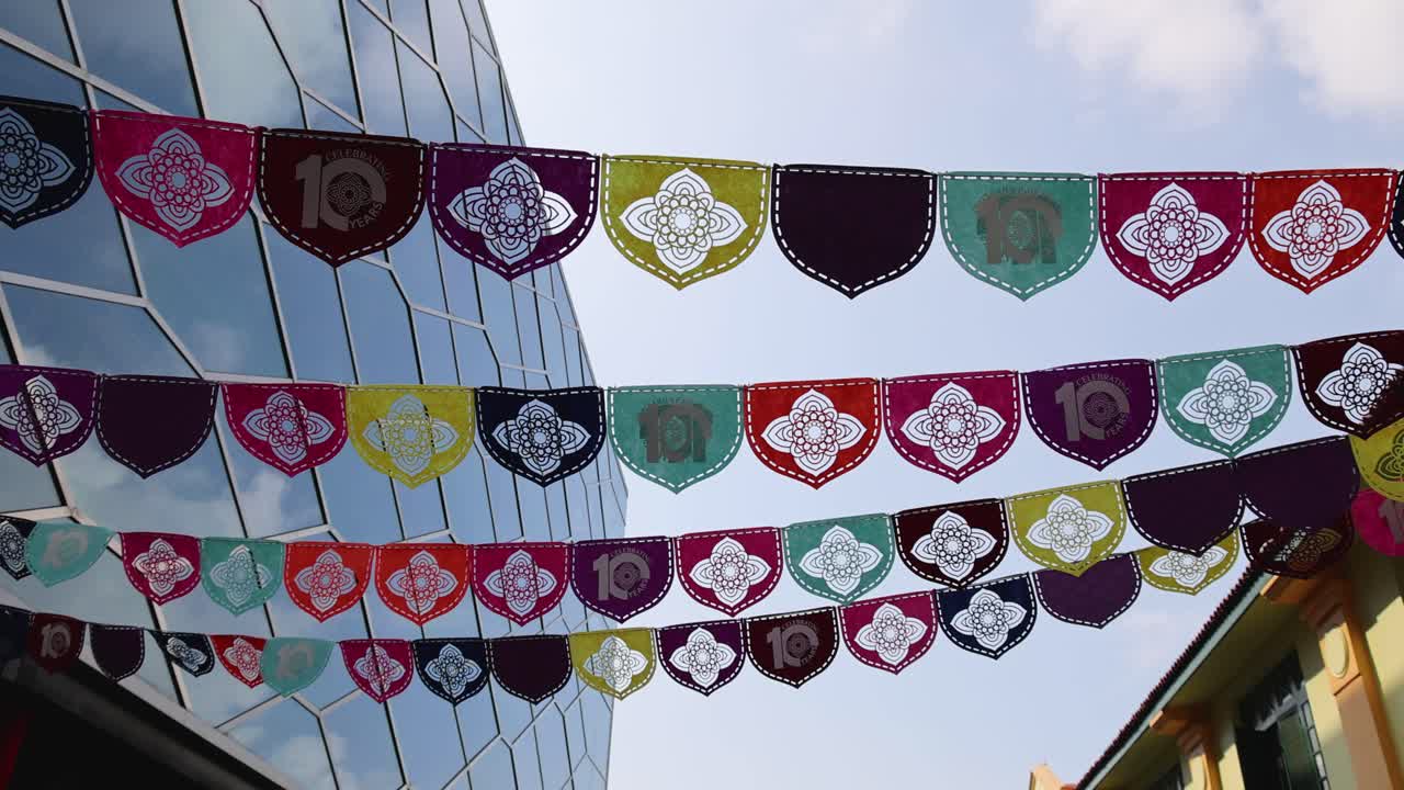 Rows of vibrant mandala paper flags flutter between modern glass buildings in Little India, Singapore, under daylight with gentle camera panning upward