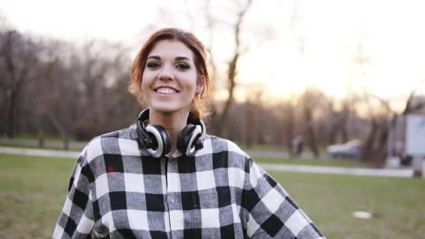 A-beautiful,-self-confident-girl-with-headphones-walks-through-the-park-in-a-good-mood.-Spinning,-posing,-looking-at-the-camera.-Casual