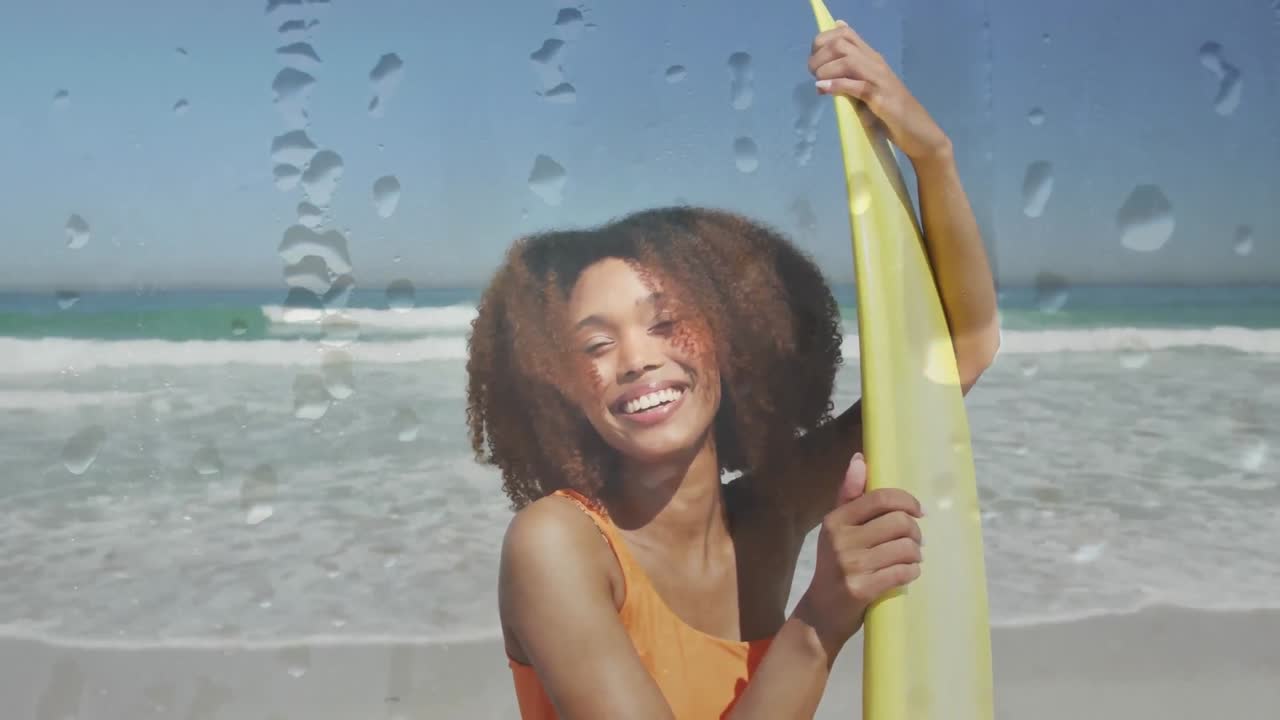 Animation of water droplets over smiling african american woman at beach with surfboard over sea