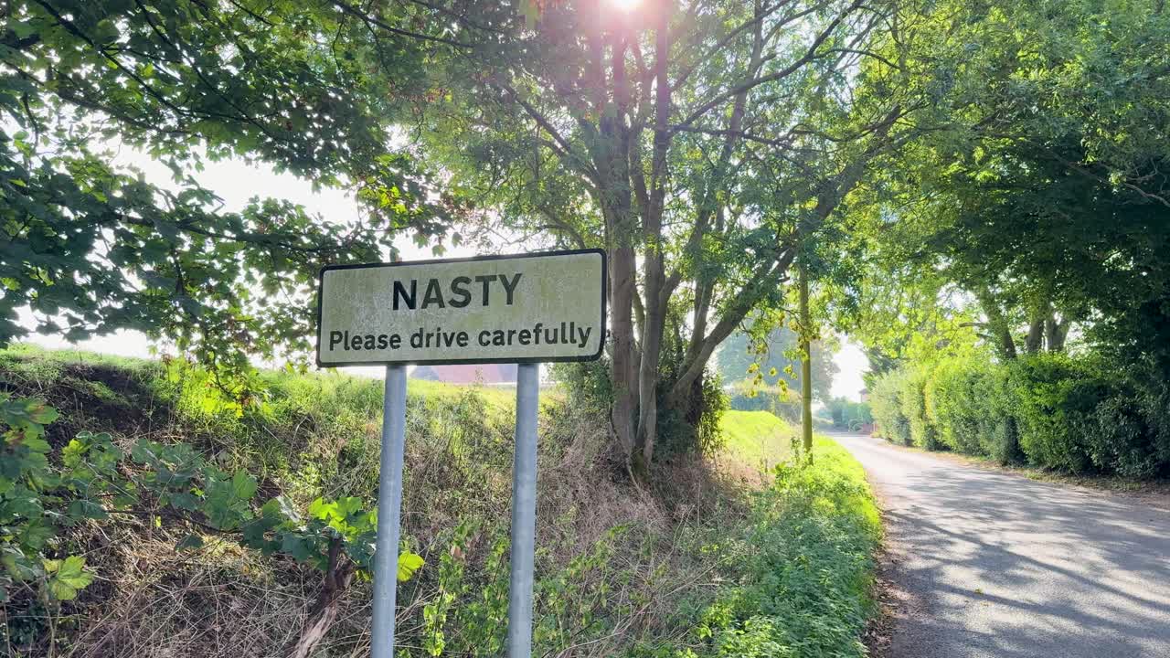 Slow-motion pan of Nasty, Hertfordshire road sign. “Please drive carefully” is visible underneath, offering both a literal traffic warning and a playful or metaphorical concept for risk and caution