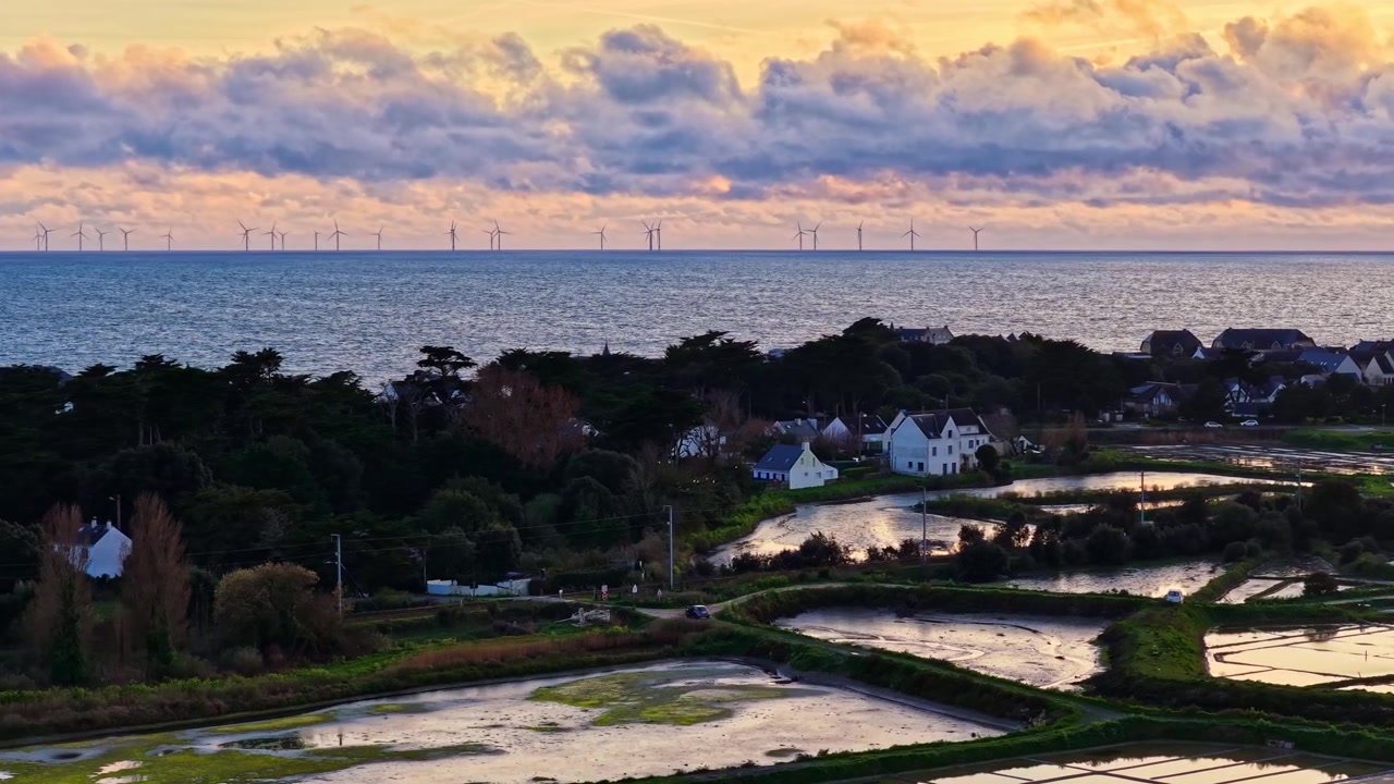 Un sereno villaggio costiero con stagni riflettenti e pale eoliche distanti sotto un cielo vibrante all'alba. Ideale per documentari ambientali o servizi di viaggio.