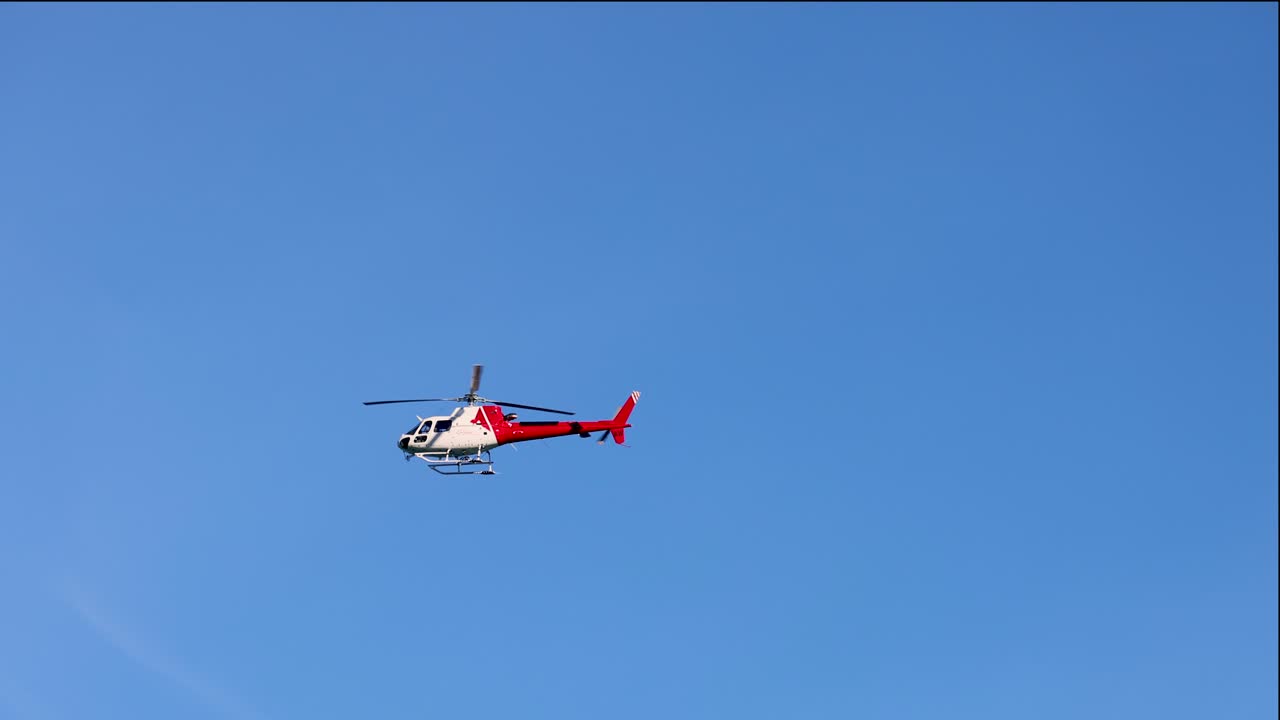 A red helicopter flies smoothly across a bright blue sky in Queenstown, New Zealand, showcasing dynamic aerial movement and serene atmosphere