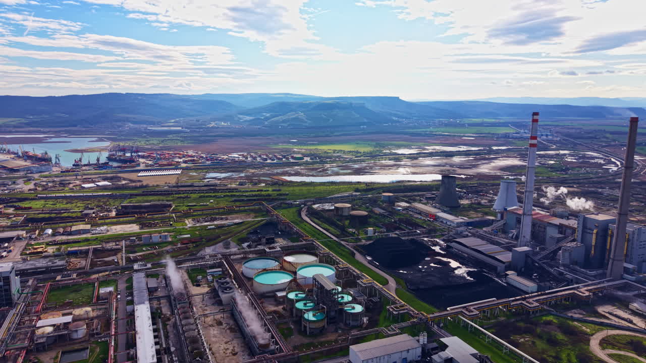 Power plant and industrial area near a river with green fields and mountains in the background during daytime