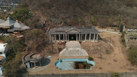 Abandoned-Greek-style-column-buildings-and-empty-blue-tile-swimming-pool-aerial-descending-view