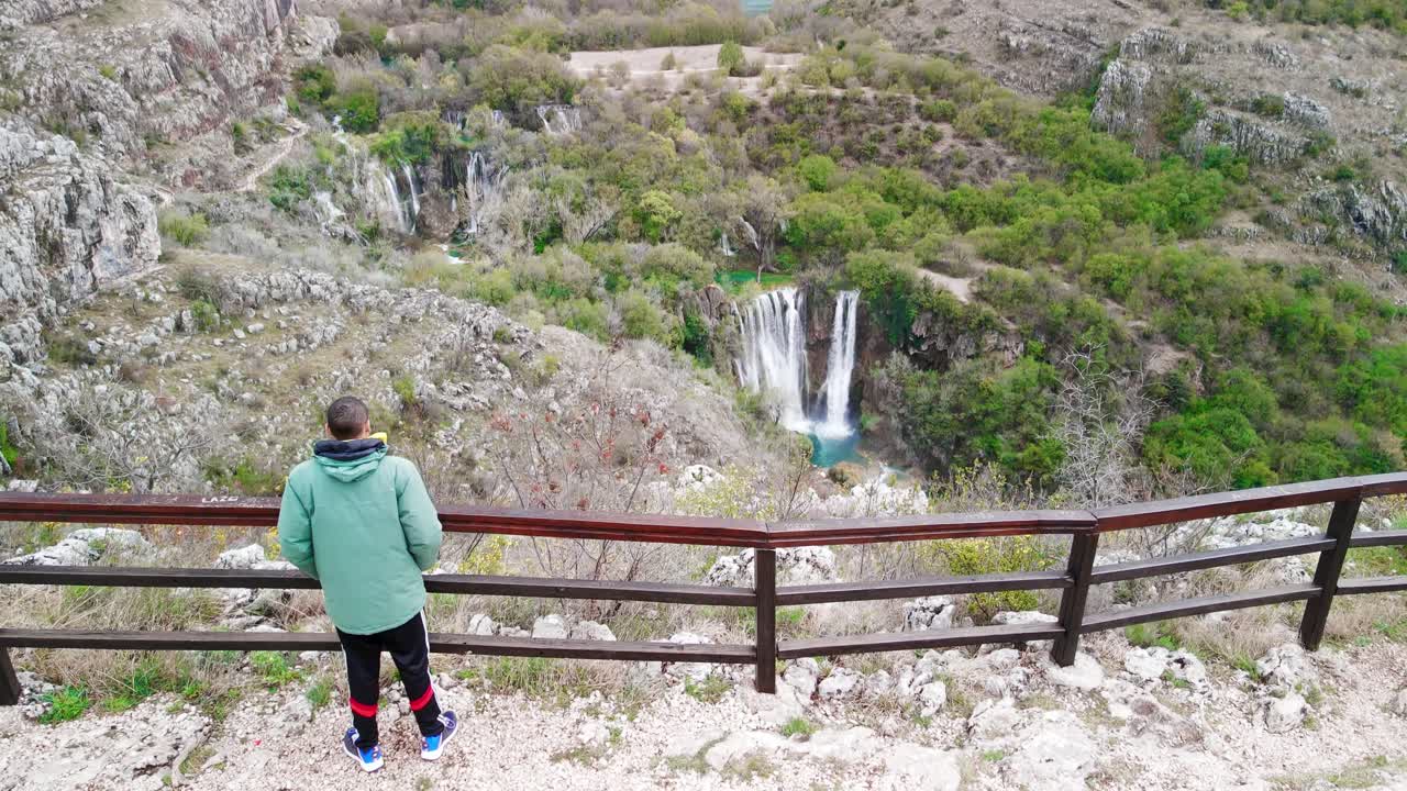 A rising and tilting aerial shot of a traveler looking out over beautiful sloping gem-toned rivers and waterfalls, and the lush terraced land around them in Krka National Park in southern Croatia