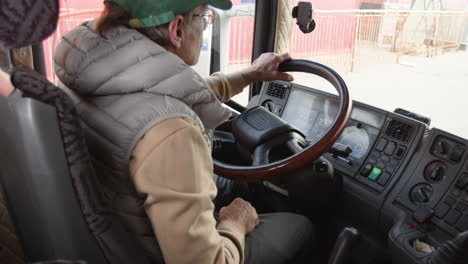 rear view of older worker wearing cap and vest driving a truck in a logistics park