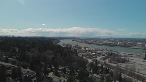 Vista-Escénica-Del-Delta-Bc-En-El-Borde-Del-Río-Fraser-Con-El-Puente-Alex-Fraser-En-El-Fondo-Día-Brillante-Cielo-Azul-Nubes-Antena-Amplia-Marcha-Atrás-Revelando-Casas-Barrio-Abajo