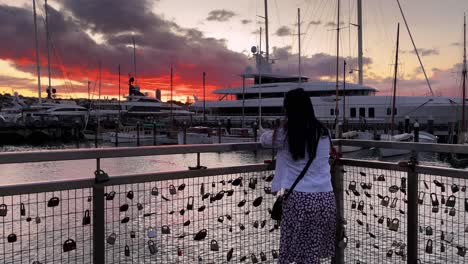 A-woman-standing-at-a-fence-with-locks-on-it-stares-at-the-sky,-water,-and-boats-in-a-marina-at-sunset