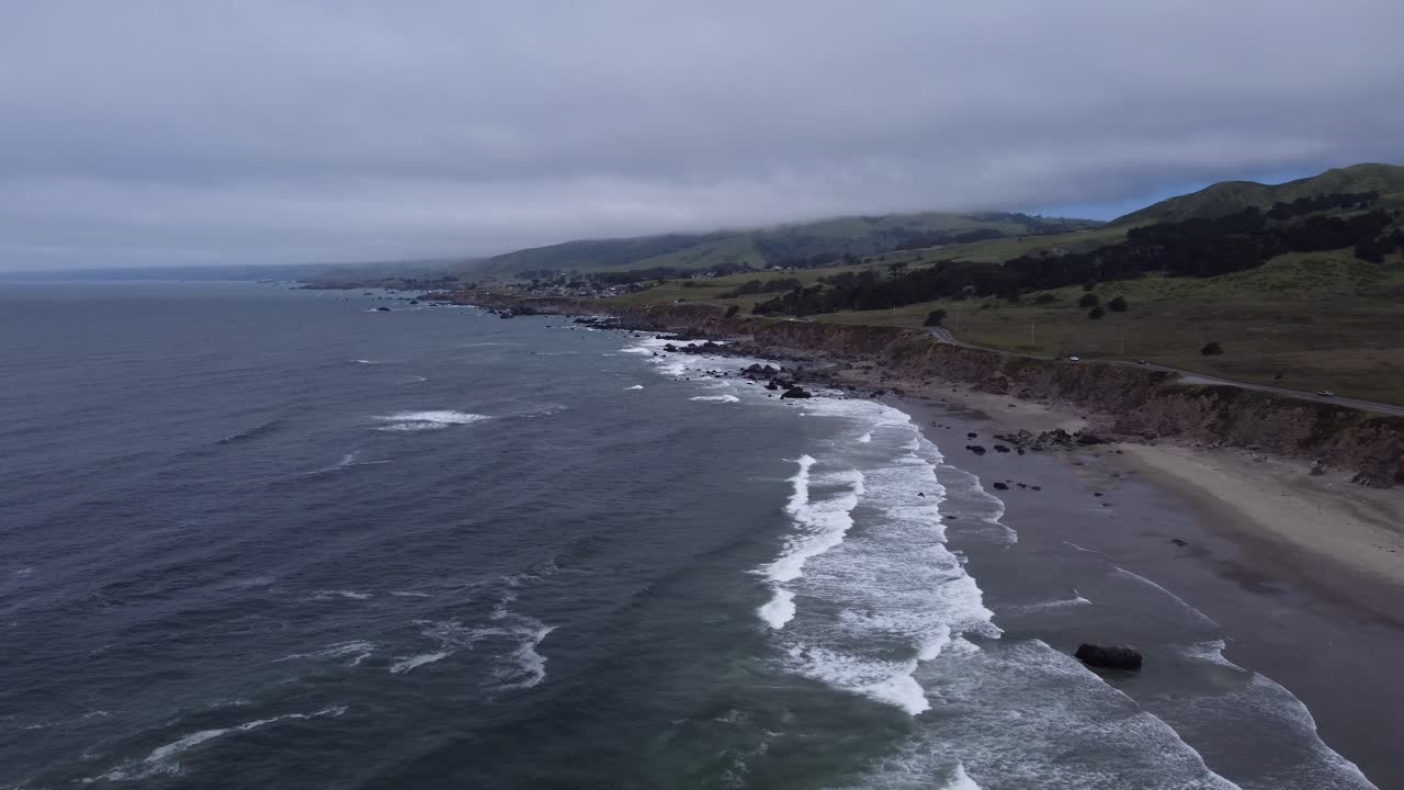 Ocean waves rolling into the Northern California coastline on a cloudy day. As the salt water batters the beach, lush green hills dotted with trees dominate the background. 4K cinematic drone footage.