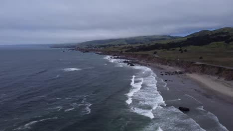 ocean waves rolling into the northern california coastline on a cloudy day. as the salt water batters the beach, lush green hills dotted with trees dominate the background. 4k cinematic drone footage.