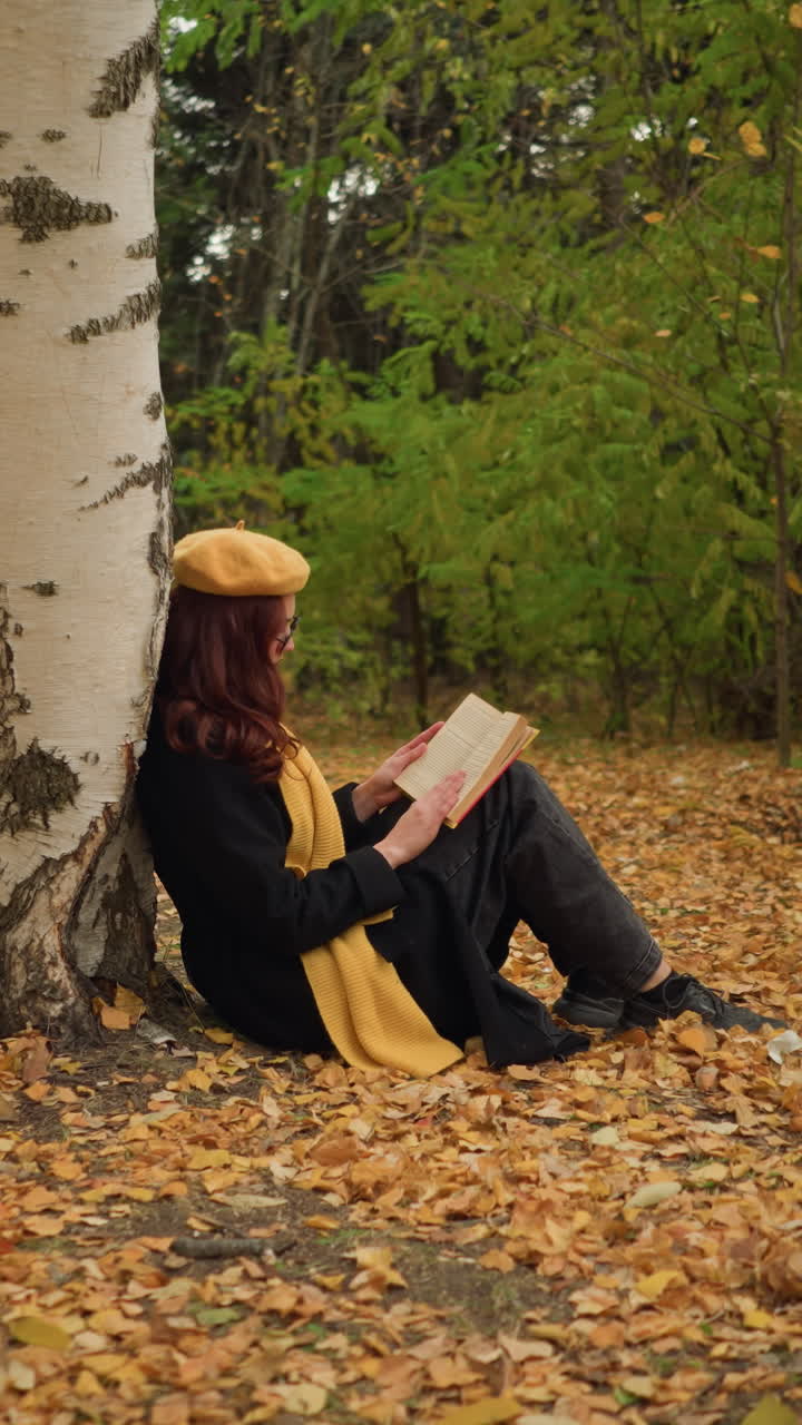 mujer vestida con trajes elegantes se sienta sola apoyándose en un árbol, leyendo un libro mientras toca suavemente las páginas, rodeada de hojas doradas de otoño en un entorno pacífico de bosque