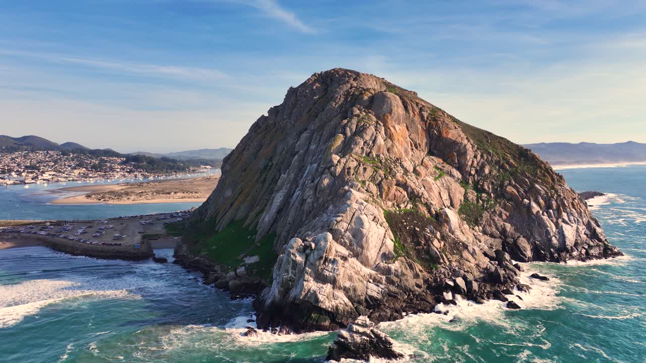 Drone establishing orbit of Morro Rock and coastal waves in Morro Bay, California under sunny skies