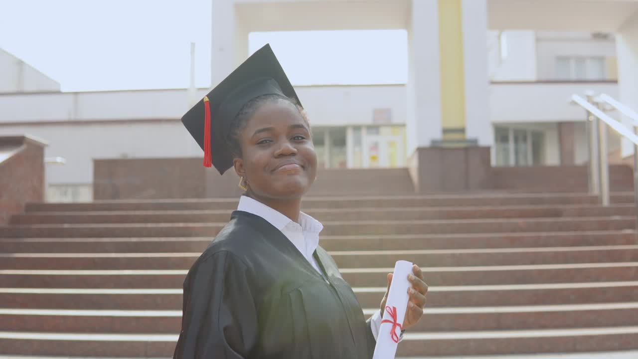joven graduada afroamericana de pie frente a la cámara con un diploma y libros en las manos. la estudiante está vestida con una túnica negra y un sombrero de maestro cuadrado y está de pie afuera.