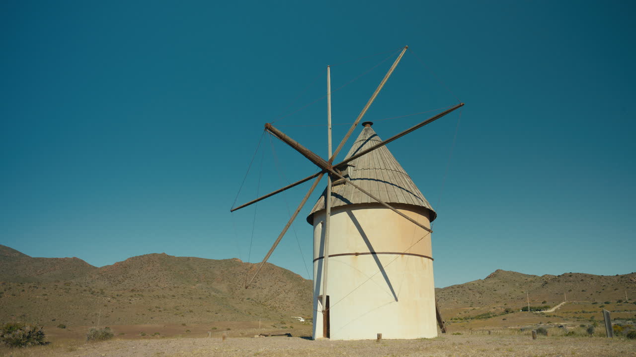 Toma de alejamiento lenta de un molino de viento tradicional en un paisaje mediterráneo seco bajo un cielo azul claro. Movimiento cinematográfico que revela las colinas circundantes y el paisaje rural