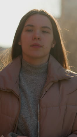 front view of a girl walking through an urban area at sunset, wearing a peach jacket and a grey inner shirt. her mouth is slightly open, and she gazes forward with a calm expression