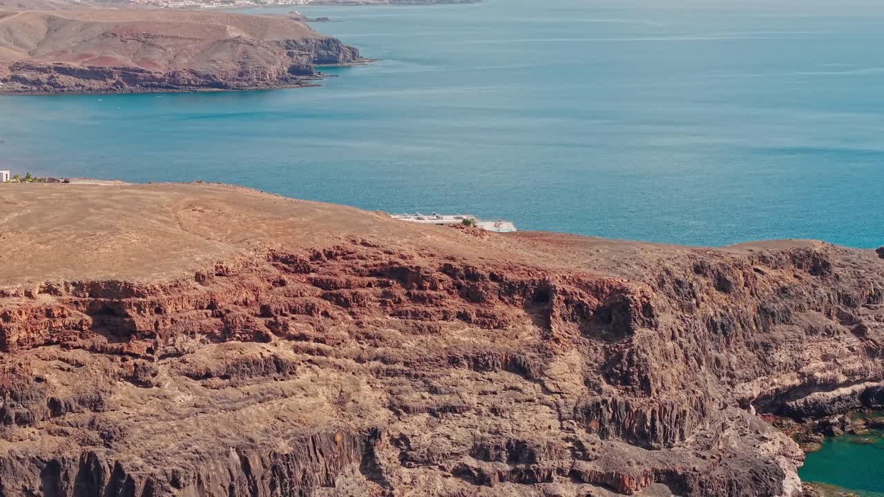 Panoramic tilt down aerial view of a hotel complex on Fuerteventura’s volcanic coastline at sunset, surrounded by warm ocean light and arid terrain, evoking calm and scenic coastal charm