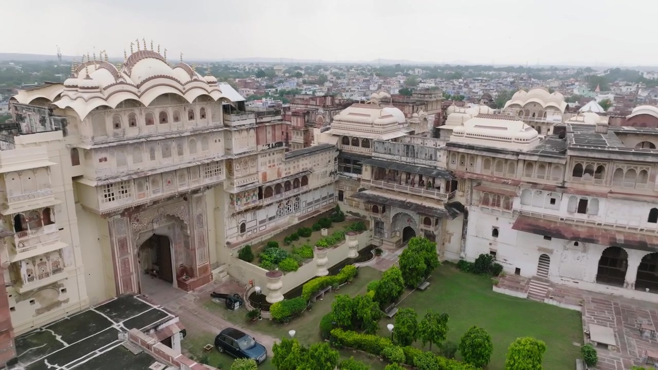 Il cortile del palazzo presenta un giardino centrale con alberi circondato da edifici storici con tetti a cupola e facciate ad arco. Una ripresa aerea con drone si sposta lentamente a destra rivelando il complesso architettonico.