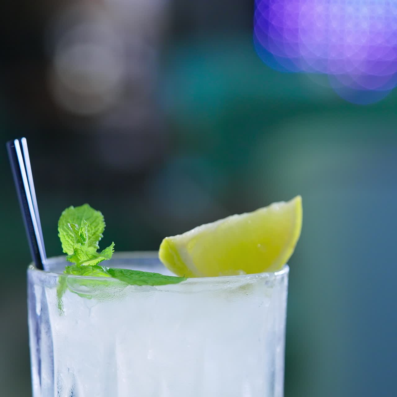 Upper part of glass filled with light beverage and two black straws in. Ice cubes, lemon slice and leaves of mint on top. Close up. Blurred backdrop