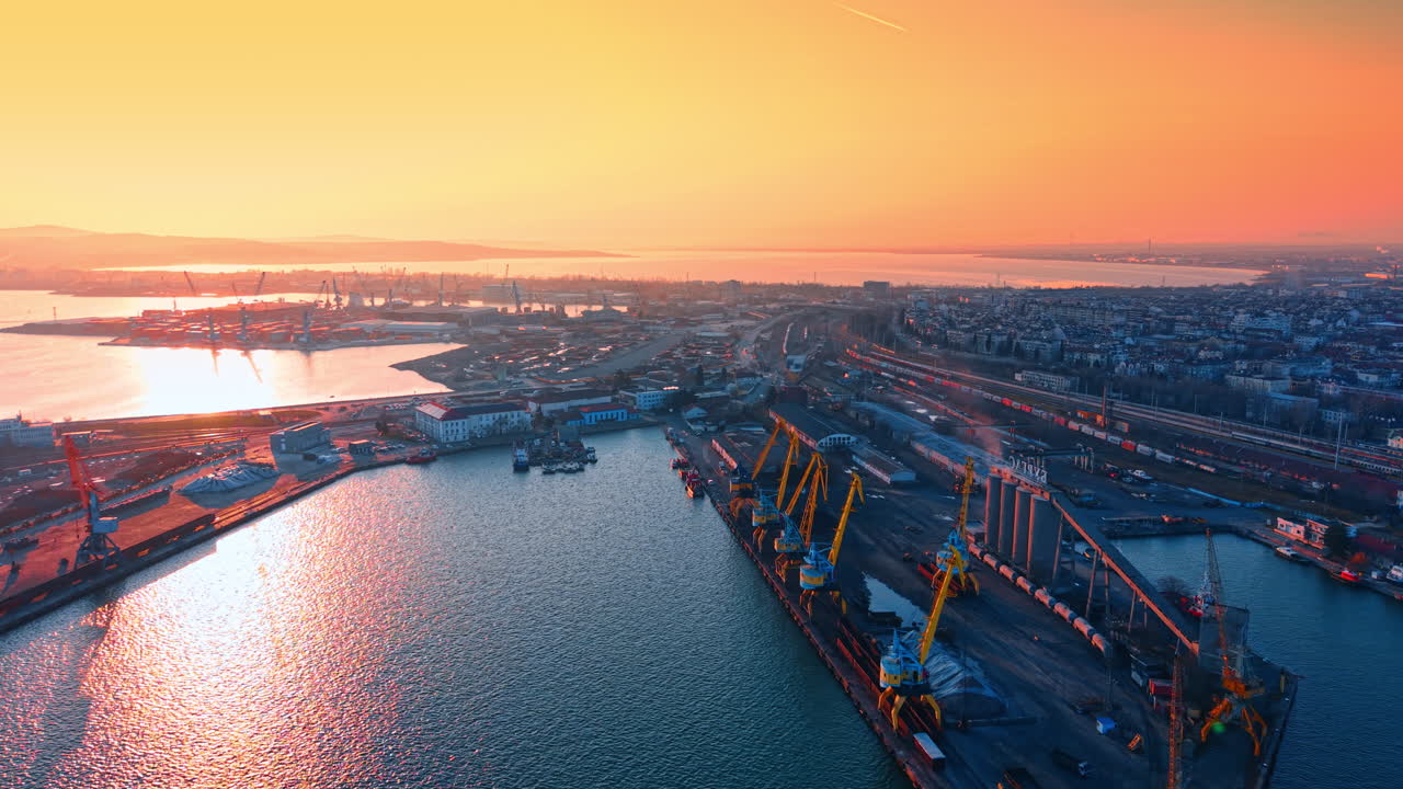 Panoramic view of sea cargo port and city. Elevated shot of industrial harbor, shipping cranes, and urban buildings under an orange sunset sky