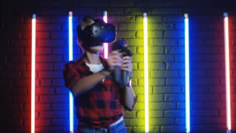 young woman in vr glasses and using joystick while playing a virtual reality game in a room with colorful neon lamps on the wall