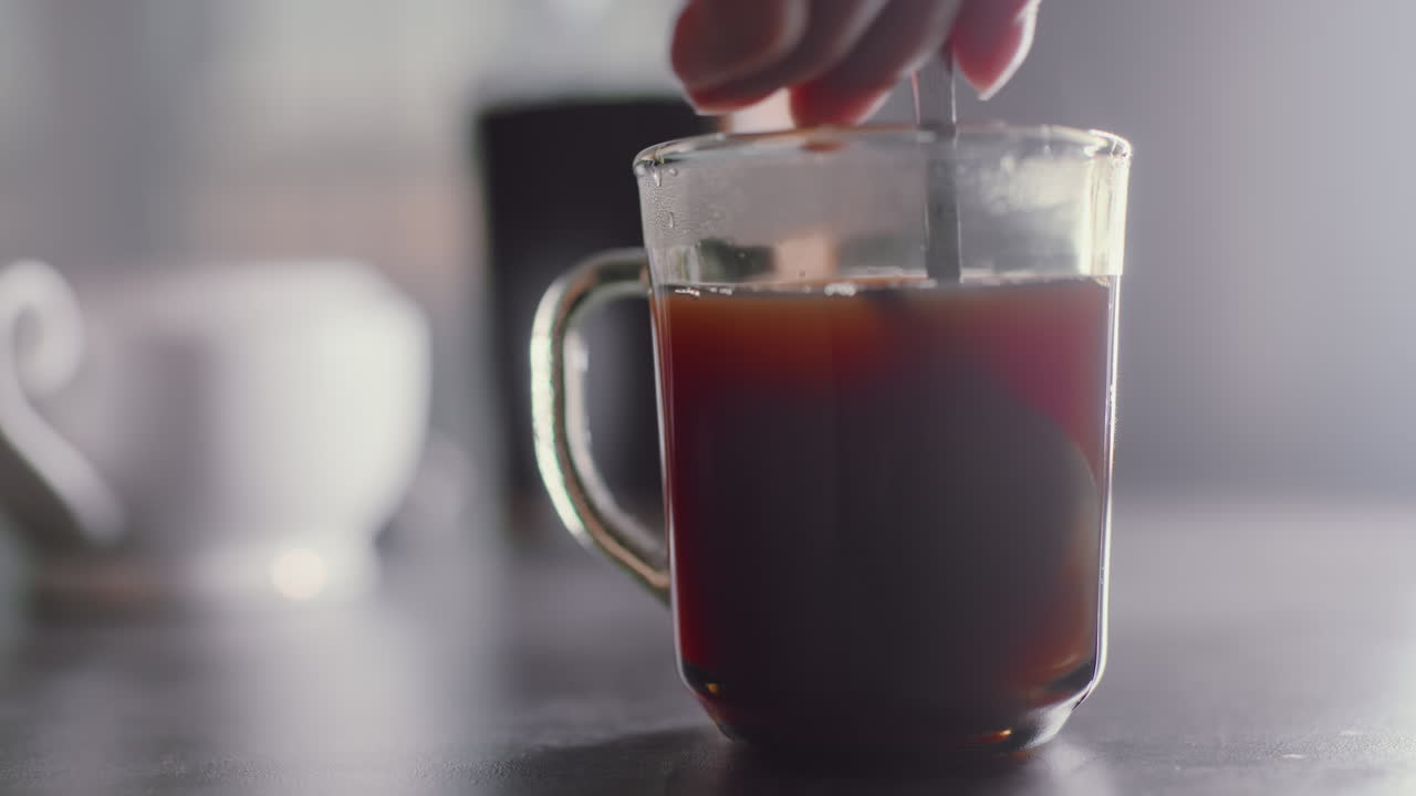 Close up of individual hand stirring coffee inside transparent glass mug during breakfast preparation, spoon swirling through steaming drink on table surface with blurred background items visible