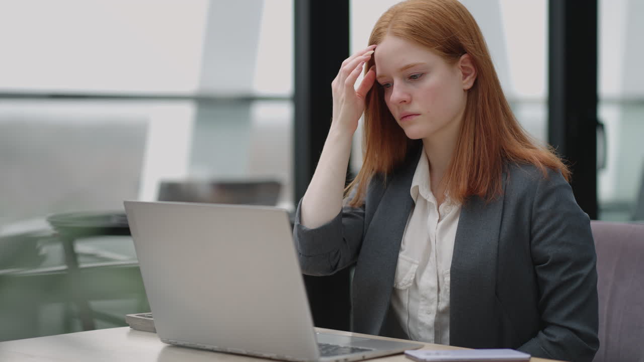 A young red-haired business woman looks thoughtfully at the screen and brainstorms. Watch and think about problems looking out the window. Thoughtful business woman