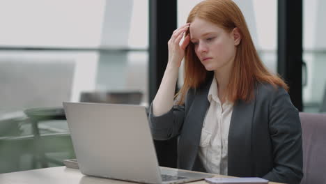 A-young-red-haired-business-woman-looks-thoughtfully-at-the-screen-and-brainstorms.-Watch-and-think-about-problems-looking-out-the-window.-Thoughtful-business-woman