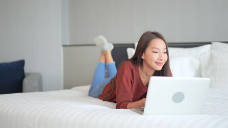 a young woman works on her laptop as she lays on her stomach on a big comfortable hotel bed