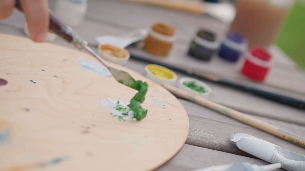 palette di legno da esterno con vernice verde, spatola che raschia una piccola quantità di vernice accanto a barattoli e pennelli su una panca scolorita, profondità di campo ridotta e luce diurna soffusa, schizzo urbano plein air