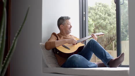 hombre bi-racial feliz sentado junto a la ventana abierta tocando la guitarra en casa, espacio de copia, cámara lenta