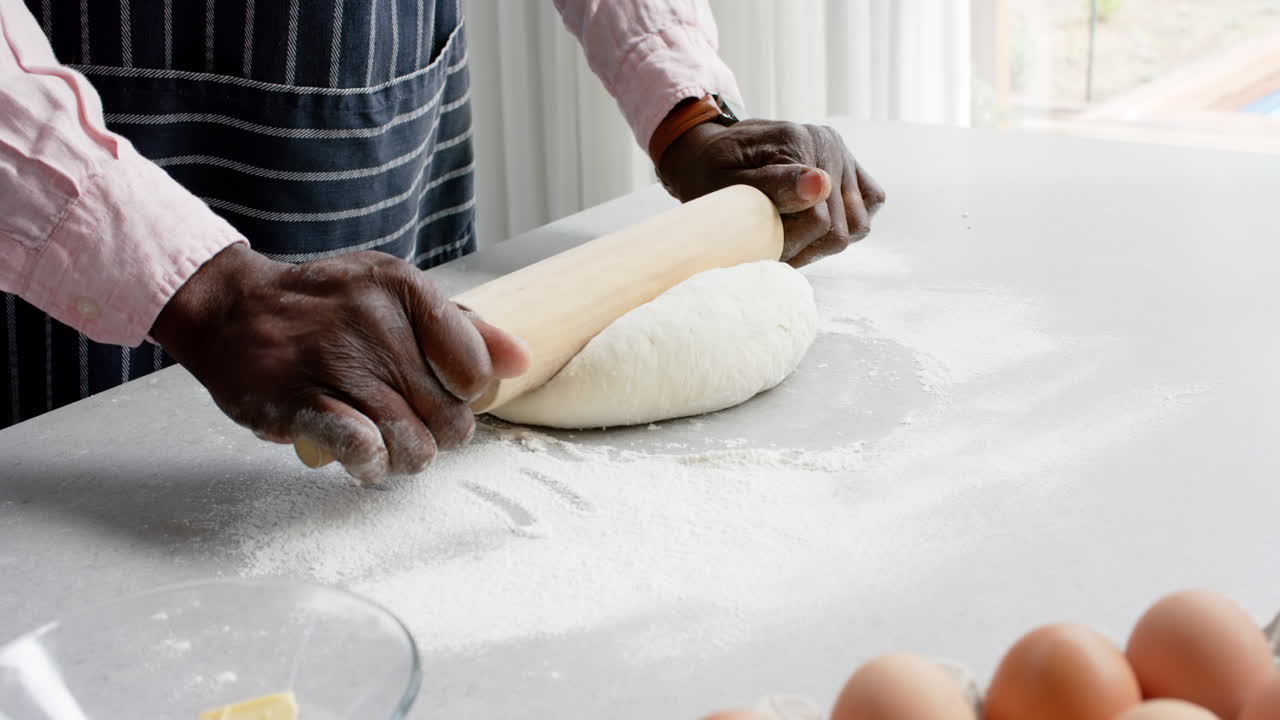 Biracial man wearing apron baking and rolling dough in kitchen, slow motion